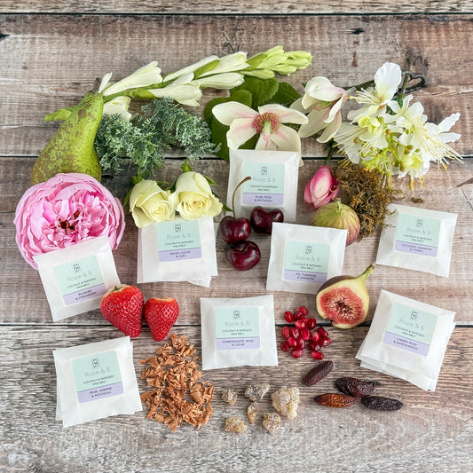 A collection of wax melt packets, each with a different fragrance, displayed alongside relevant botanicals such as flowers and fruits, on a wooden surface.